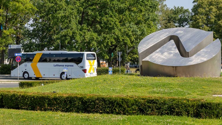 Nuremberg Airport, August 4, 2026 A Lufthansa Express tour bus passes by a distinctive metal logo at a roundabout at the entrance to the airport grounds. In the background, a sign pointing to the airport can be seen. Nuremberg Airport, Bavaria, Germany 20260804-1V9A6673-M4000