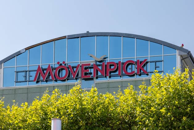 Nuremberg Airport, August 4, 2026 A Mövenpick Hotel building with a distinctive logo directly across from the airport terminal The curved glass facade reflects the blue sky Nuremberg Airport, Bavaria, Germany 20260804-1V9A6688-M4000