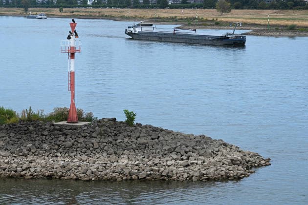 Historically low water levels on the Rhine; the Düsseldorf Water Police report that the Rhine’s water level in Düsseldorf has reached 23 cm—the lowest ever recorded there. Low water level, rocky riverbank, a tall red bank marker, and a cargo ship in the background, Düsseldorf, August 4, 2026