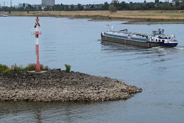 Historically low water levels on the Rhine; the Düsseldorf Water Police report that the Rhine’s water level in Düsseldorf has reached 23 cm—the lowest ever recorded there. Low water level, rocky riverbank, a tall red bank marker, and a cargo ship in the background, Düsseldorf, August 4, 2026