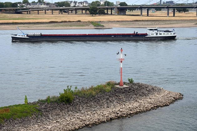 Historically low water levels on the Rhine; the Düsseldorf Water Police report that the Rhine’s water level in Düsseldorf has reached 23 cm—the lowest ever recorded there. Low water level, rocky riverbank, a tall red bank marker, and a cargo ship in the background, Düsseldorf, August 4, 2026
