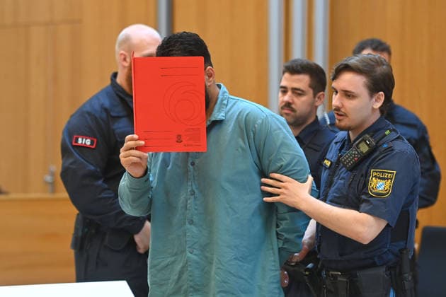 The defendant is escorted into the courtroom by court officers. The verdict in the criminal case against Farhad Noori for murder, among other charges, related to the Seidlstrasse incident will be handed down on August 6, 2026, at the Munich Higher Regional Court.