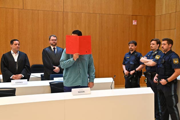 The defendant in the courtroom hides his face behind a binder. Behind him are his defense attorneys, Oemer SAHINCI, Esq., and Johann BUND, Esq. Judgment in the criminal case against Farhad Noori for murder, among other charges, on Seidlstrasse on August 6, 2026, before the Munich Higher Regional Court