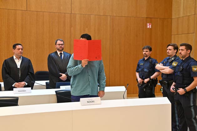 The defendant in the courtroom hides his face behind a binder. Behind him are his defense attorneys, Oemer SAHINCI, Esq., and Johann BUND, Esq. Judgment in the criminal case against Farhad Noori for murder, among other charges, on Seidlstrasse on August 6, 2026, before the Munich Higher Regional Court