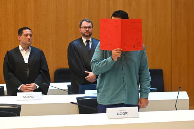 The defendant in the courtroom hides his face behind a binder. Behind him are his defense attorneys, Oemer SAHINCI, Esq., and Johann BUND, Esq. Judgment in the criminal case against Farhad Noori for murder, among other charges, on Seidlstrasse on August 6, 2026, before the Munich Higher Regional Court