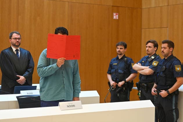 The defendant in the courtroom hides his face behind a binder. Behind him is his defense attorney, Johann BUND. Judgment in the criminal case against Farhad Noori for murder, among other charges, on Seidlstrasse on August 6, 2026, before the Munich Higher Regional Court.