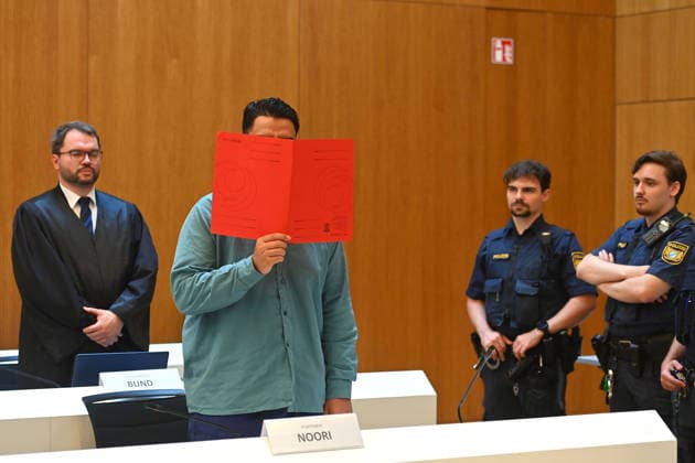 The defendant in the courtroom hides his face behind a binder; behind him stands his defense attorney, Johann BUND. Verdict in the criminal case against Farhad Noori for murder, among other charges, on Seidlstrasse on August 6, 2026, before the Munich Higher Regional Court.