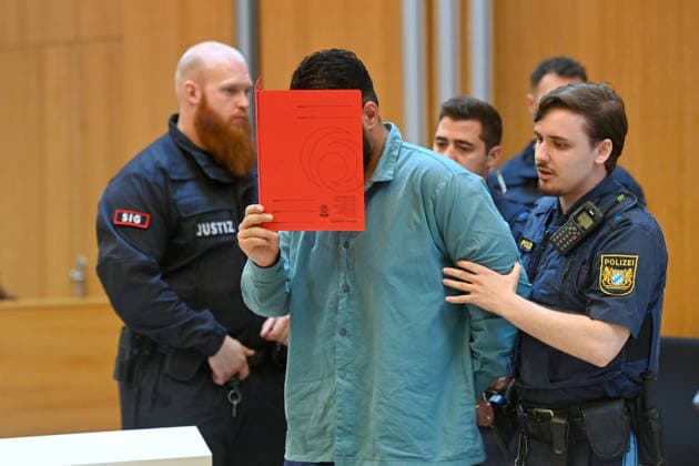 The defendant is escorted into the courtroom by court officers. The verdict in the criminal case against Farhad Noori for murder, among other charges, related to the Seidlstrasse incident will be handed down on August 6, 2026, at the Munich Higher Regional Court.