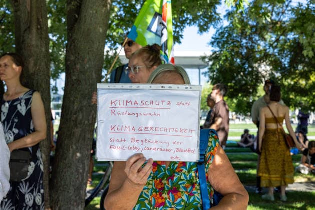 Fridays for Future Berlin Heat Protest, Germany, Berlin, August 6, 2026. A woman holds up a small sign reading: Climate Protection Instead of the Arms Race; Climate Justice Instead of Catering to the Fossil Fuel Lobby, Land Gr