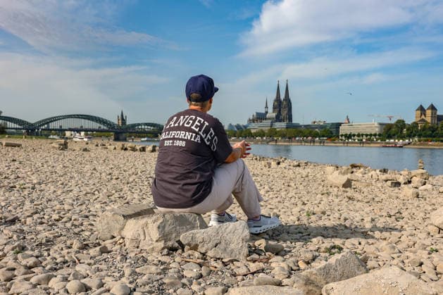 Summer in the Rhineland: View of the Rhine near Cologne (Deutz). The Rhine in Cologne is at low water levels due to persistent drought. In the background, the Hohenzollern Bridge is on the left and Cologne Cathedral is on the right. In the foreground, a woman is sitting on a rock, looking out at the Rhine. Summer in the Rhineland on Augus