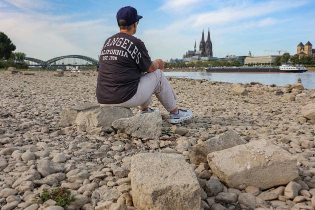Summer in the Rhineland: View of the Rhine near Cologne (Deutz). The Rhine in Cologne is at low water levels due to persistent drought. In the background, the Hohenzollern Bridge is on the left and Cologne Cathedral is on the right. In the foreground, a woman is sitting on a rock, looking out at the Rhine. Summer in the Rhineland on Augus