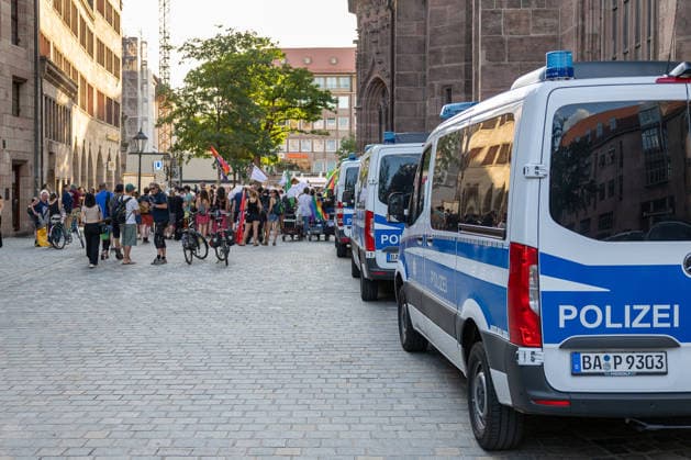 Police operation during the Monday demonstration by Team Menschenrechte and counter-protests, Nuremberg, 10 087 2026 Several police vehicles are parked on the east side of St. Lorenz Church, while a group of demonstrators carrying flags and banners gathers. Passersby on bicycles watch the scene unfold on the cobblestone street. Nuremberg, Bavaria, Germany 20260810-6V2A0609-M4000