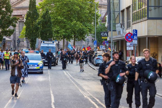 Police operation during the Monday demonstration by Team Menschenrechte and counter-protests, Nuremberg, 10 087 2026 Police officers in protective gear escort a demonstration march carrying flags through a downtown street. Police vehicles secure the procession, while passersby watch the events unfold from the sidewalk. Nuremberg, Bavaria, Germany 20260810-286A5031-M4000