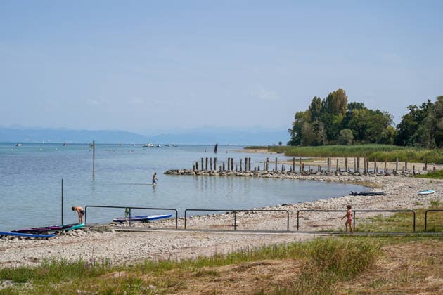 Switzerland, Lake Constance: Low water levels expose wide stretches of gravel and shoreline; swimmers stand in shallow water, SUP boards lie on the shore in the background; boats and an Alpine panorama. Stock image representing Lake Constance, water level, low water, drought, heat, heat wave, summer, climate change, bodies of water, shore, gravel beach Copyright: xJOERANxSTEINSIEKx