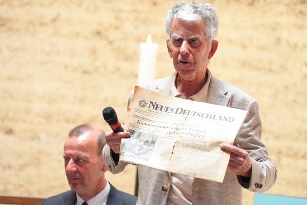 Commemoration of the 65th Anniversary of the Construction of the Berlin Wall: Jörg Hildebrandt holds up the historic daily newspaper *Neues Deutschland* and its front page, pointing to the headline about communism at the commemoration marking the 65th anniversary of the construction of the Berlin Wall, Berlin, August 13, 2026 Berlin Berlin Germany