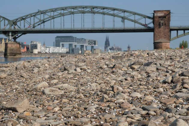 A low-angle shot captures the wide, exposed pebble and stone bank of the Rhine during extreme summer low water levels. In the blurred background, the steel truss structure of the historic Südbrücke stands out with its distinctive brick defense towers. Behind it, the Cologne skyline features the 20260816-DSCF2111