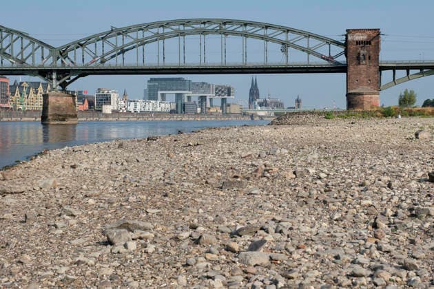 A low-angle shot captures the wide, exposed pebble and stone bank of the Rhine during extreme summer low water levels. In the blurred background, the steel truss structure of the historic Südbrücke stands out with its distinctive brick defense towers. Behind it, the Cologne skyline fe 20260816-DSCF2116