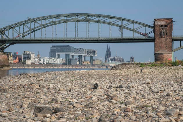 A low-angle shot captures the wide, exposed pebble and stone bank of the Rhine during extreme summer low water levels. In the blurred background, the steel truss structure of the historic Südbrücke stands out with its distinctive brick defense towers. Behind it, the Cologne skyline fe 20260816-DSCF2120