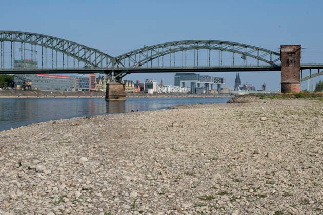 A low-angle shot captures the wide, exposed pebble and stone bank of the Rhine during extreme summer low water levels. In the blurred background, the steel truss structure of the historic Südbrücke stands out with its distinctive brick defense towers. Behind it, the Cologne skyline fe 20260816-DSCF2122