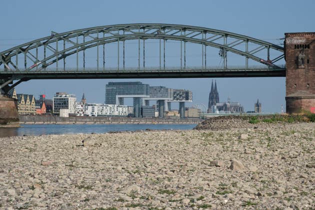A low-angle shot captures the wide, exposed pebble and stone bank of the Rhine during extreme summer low water levels. In the blurred background, the steel truss structure of the historic Südbrücke stands out with its distinctive brick defense towers. Behind it, the Cologne skyline features the 20260816-DSCF2123