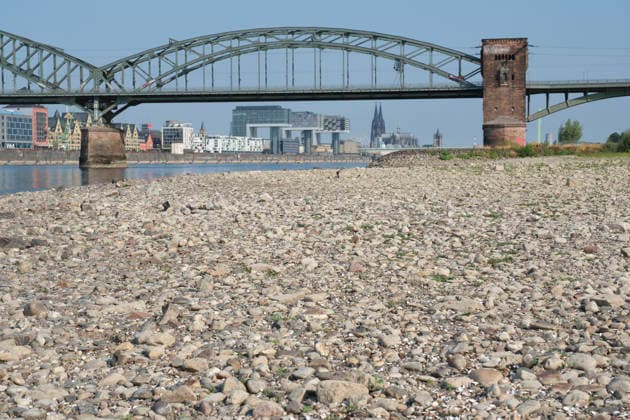 A low-angle shot captures the wide, exposed pebble and stone bank of the Rhine during extreme summer low water levels. In the blurred background, the steel truss structure of the historic Südbrücke stands out with its distinctive brick defense towers. Behind it, the Cologne skyline features the 20260816-DSCF2126