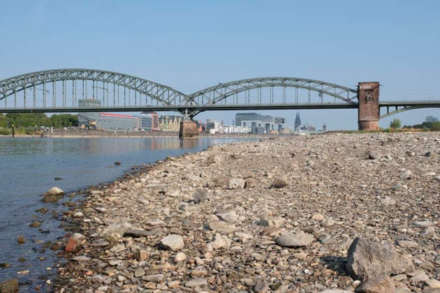 A wide-angle view captures the significantly receded riverbed of the Rhine, exposing a vast dried-out pebble and stone bank during extreme summer low water levels. The historic steel Südbrücke spans the river with 20260816-DSCF2131