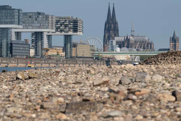 A telephoto shot captures the prominent Cologne cityscape featuring the modern Kranhäuser buildings in the Rheinauhafen and the Gothic spires of the Cologne Cathedral in the background. In the blurred foreground, the vast, exposed pebble and stone bank of the Rhine illustrates the impact of extreme summer low water levels. Next to the cathedral, the white Nordrhein-Westfalen Deutschland, Germany 20260816-DSCF2154