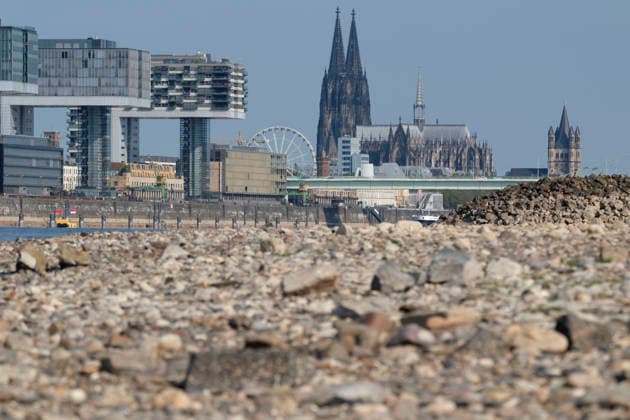 A telephoto shot captures the prominent Cologne cityscape featuring the modern Kranhäuser buildings in the Rheinauhafen and the Gothic spires of the Cologne Cathedral in the background. In the blurred foreground, the vast, exposed pebble and stone bank of the Rhine illustrates the impact of extreme summer low water levels. Next to the cathedral, the white Nordrhein-Westfalen Deutschland, Germany 20260816-DSCF2155
