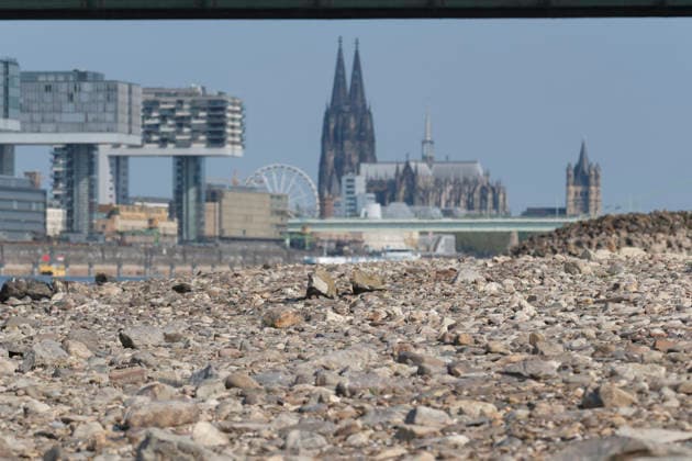 A telephoto shot captures the prominent Cologne cityscape featuring the modern Kranhäuser buildings in the Rheinauhafen and the Gothic spires of the Cologne Cathedral in the background. In the foreground, the vast, exposed pebble and stone bank of the Rhine illustrates the impact of extreme summer low water levels. Next to the cathedral, the white Nordrhein-Westfalen Deutschland, Germany 20260816-DSCF2159