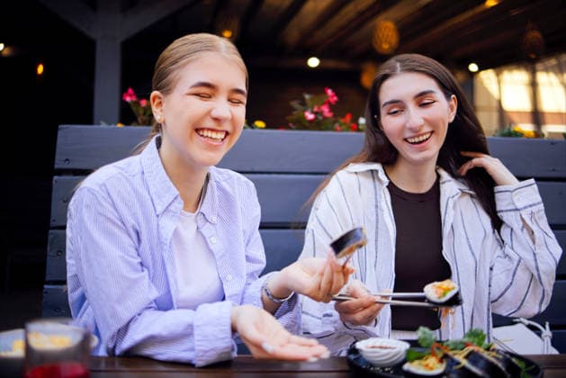 Zwei junge Frauen genießen Sushi in einem Restaurant im Freien, lächeln und lachen, während sie gemeinsam essen; im Hintergrund sind bunte Blumen und eine Holzdekoration zu sehen Two smiling young women share sushi at an outdoor dining table, surrounded by flowers and wooden decor, creating a lively dining atmosphere (License=RF) ,model released, Symbolfoto Copyright: xZoonar.com DashaxPetrenkox 25530421 ,model released, Symbolfoto ,property released