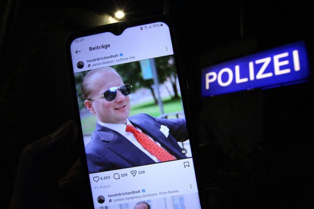 A man is standing in front of a police station holding a smartphone, on which the Instagram account of wind power fraudster Hendrik Holt is open. After being granted a temporary release, he failed to return to the correctional facility in Ber