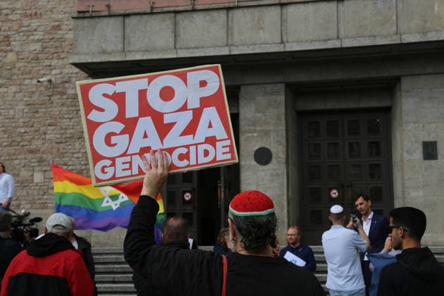 Berlin, Germany, September 17, 2026: A man holds a sign reading STOP GAZA GENOCIDE in front of Neukölln City Hall during the Berlin Wears the Star of David solidarity rally. In the background are participants in the rally and a rainbow flag featuring the Star of David. Gaza, Israel, Middle East conflict, protest, counter-protest, Star of David, Copyright: xAlonxDavidx