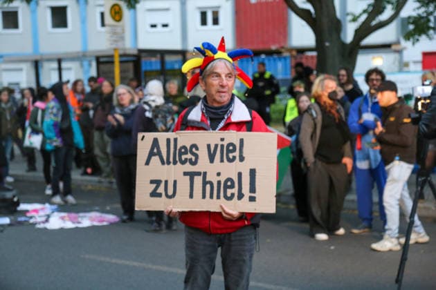 Berlin, Germany, September 24, 2026: A participant in the protest rally against the presentation of the Axel Springer Award to Peter Thiel holds a sign reading Everything’s Too Much for Thiel and wears a colorful jester’s cap. In the background are demonstrators and a Palestinian flag. Demonstration, protest, Peter Copyright: xAlonxDavidx
