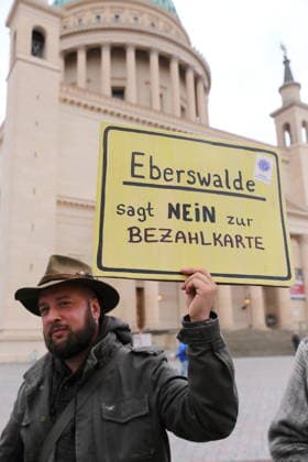 A protester stands outside the state parliament holding a sign that reads Eberswalde Says No to the Payment Card during a demonstration against the payment card for refugees in Brandenburg, Potsdam, September 25, 2026. Demonstration against the payment card in Potsdam