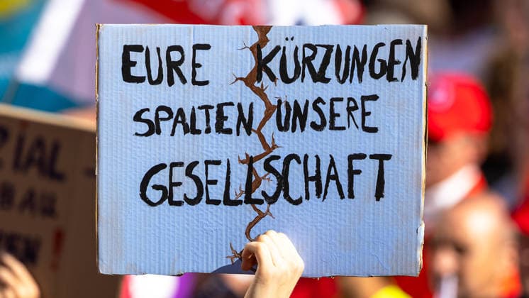 DGB Bavaria: Hard-Earned, Big Rally, Nuremberg, September 26, 2026 A protester holds aloft a cardboard sign reading Your cuts are dividing our society. A painted crack runs symbolically through the center of the sign; the hand holds it firmly. Nuremberg, Bavaria, Germany 20260926-1V9A9949-M4000