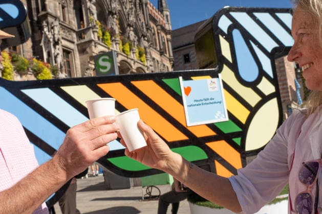 Münchner Olympia-Bewerbung erfolgreich, München wird deutscher Olympia-Bewerber, Münchner stoßen darauf an und auf einen Geburtstag, Olympia-Maskottchen Dackel Waldi auf dem Marienplatz, München, 26. September 2026 Deutschland, München, 26. September 2026, Münchner Olympia-Bewerbung erfolgreich, München wird deutscher Olympia-Bewerber, Mitglieder einer kleinen Geburtstagsparty stoßen auf Geburtstag und die Olympia-Entscheidung an, Sekt aus Pappbechern, Olympia-Maskottchen Dackel Waldi auf dem Marienplatz, Münchner Olympia-Bewerbung mit drei Meter hohem Riesen-Dackel, Schild: Heute fällt die Entscheidung, Marienplatz, Entscheidung des Deutschen Olympischen Sportbunds DOSB für München als Austragungsort der Olympiade 2036, 2040 o