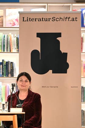 Minifestival AH0J (Literaturfestival) - Tschechien setzt über - der Kulturinitiative Literaturschiff, in der Buchhandlung Kurdirektion in Bad Ischl. Lesungen und Gespräche in Korrespondenz zum Gastauftritt Tschechiens auf der Frankfurter Buchmesse 2026. Es liest die tschechische Schriftstellerin und mehrfache Literaturpreisträgerin Radka Denemarkovich. Die Veranstaltung wrd moderiert von Julia Miesenböck, fotografiert am 30.09.26. Das Bild zei vor einem Plakat der Kulturinitiative Literaturschiffgt die Schriftstellerin Radka Denemarkovic 2026 - Minifestival AH0J (Literaturfestival) - Tschechien setzt über - der Kulturinitiative Literaturschiff, in der Buchhandlung Kurdirektion in Bad Ischl. Lesungen und Gespräche in Korrespondenz zum