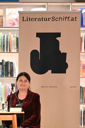 Minifestival AH0J (Literaturfestival) - Tschechien setzt über - der Kulturinitiative Literaturschiff, in der Buchhandlung Kurdirektion in Bad Ischl. Lesungen und Gespräche in Korrespondenz zum Gastauftritt Tschechiens auf der Frankfurter Buchmesse 2026. Es liest die tschechische Schriftstellerin und mehrfache Literaturpreisträgerin Radka Denemarkovich. Die Veranstaltung wrd moderiert von Julia Miesenböck, fotografiert am 30.09.26. Das Bild zei vor einem Plakat der Kulturinitiative Literaturschiffgt die Schriftstellerin Radka Denemarkovic 2026 - Minifestival AH0J (Literaturfestival) - Tschechien setzt über - der Kulturinitiative Literaturschiff, in der Buchhandlung Kurdirektion in Bad Ischl. Lesungen und Gespräche in Korrespondenz zum