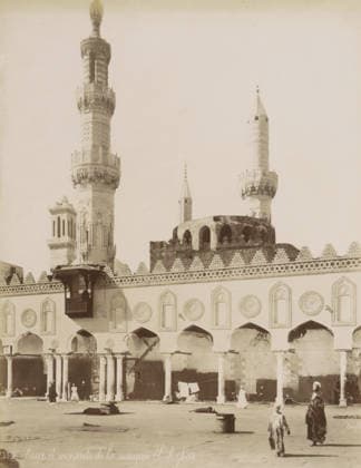RECORD DATE NOT STATED Courtyard and minarets of the Al-Azhar Mosque in Cairo, Cour et minarets de la mosquà e El-Azhar, photograph, Part of Travel Album with 80 Photos of Egypt., negative number: 1369 , photographer: Maison Bonfils, Cairo (city), c. 1870 - c. 1898, Ottoman Empire, North Africa, pasteboard, paper, albumen print, height 280 mm x width 218 mm, temple, shrine, Islam, Mohammedanism, An exterior view of a large ornate building with arched openings along its facade and decorative circular and pointed panel designs above each arch, a patterned crenellated parapet along the roofline, and tall slender towers rising behind it with carved and geometric detailing on their shafts and ornate balconies, one tower topped with a bulbous finial and another with a Copyright Penta Springs LCK260531_03292
