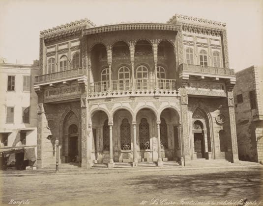RECORD DATE NOT STATED Le Caire, Fontaine de la Validà (Egypt), Building with well in Cairo, photograph, Part of Travel Album with 80 Photos of Egypt., negative number: 80 , photographer: Maison Bonfils, Cairo (city), c. 1870 - c. 1898, Ottoman Empire, North Africa, pasteboard, paper, albumen print, height 220 mm x width 281 mm, utilitarian, public pump or water fountain, A frontal view of a large ornate building facade with an upper level featuring rows of arched windows and decorative railings, and a lower level with a series of tall arches supported by slender columns leading to intricately patterned window grilles and doorways; the facade displays carved and geometric ornamentation around arches and windows, latticework balconies, and decorative friezes alo Copyright Penta Springs LCK260531_03306
