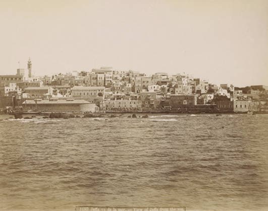 RECORD DATE NOT STATED Jaffa, seen from the sea, Jaffa vu de la mer. - View of Jaffa from the sea., photograph, Part of Travel Album with Photos of Jerusalem and Other Biblical Places., negative number: 1193 , photographer: attributed to Maison Bonfils, c. 1867 - c. 1895, Ottoman Empire, Attributed to, pasteboard, paper, albumen print, height 222 mm x width 282 mm, prospect of city, town panorama, silhouette of city, A wide waterfront view showing a dense cluster of multi-level stone and plaster buildings along a shoreline, with flat roofs, domes, arched windows and doorways, and a tall tower with a pointed top rising above the other structures, all rendered in a sepia tone; in front of the buildings a narrow quay with small boats and masts is visible, dark rock Copyright Penta Springs LCK260531_03377