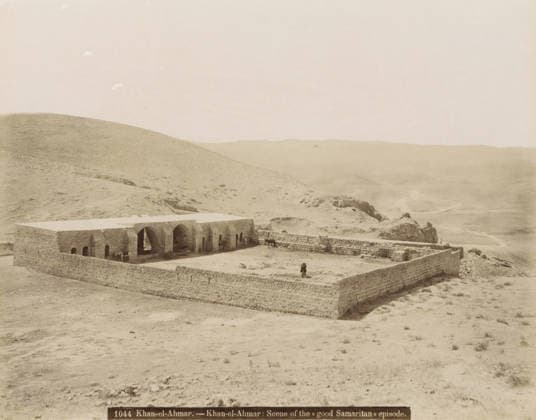 RECORD DATE NOT STATED Khan al-Ahmar.- Khan al-Ahmar: Scene of the Good Samaritan episode., Walled building in Khan al-Ahmar, photograph, Part of travel album with photos of Jerusalem and other biblical places., negative number: 1044 , photographer: attributed to Maison Bonfils, Khan Al-Ahmar, c. 1867 - c. 1895, Palestine, West Bank, Attributed to, pasteboard, paper, albumen print, height 225 mm x width 283 mm, fence, wall, paling, desert, A stone enclosure sits in a barren landscape, its perimeter formed by low rough stone walls surrounding a rectangular courtyard and a long building with a flat roof and a series of arched openings along one side; inside the courtyard and near the arches are a few animals and figures, and a person stands near the outer wall; Copyright Penta Springs LCK260531_03378