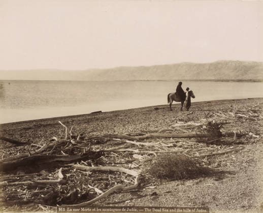 RECORD DATE NOT STATED Rider and man on the shore of the Dead Sea with washed-up branches in the foreground, La mer Morte et les montagnes de Judà e.- The Dead Sea and the hills of Judea., photograph, Part of Travel album with photos of Jerusalem and other biblical places., negative number: 911 , photographer: Maison Bonfils, Dead Sea, c. 1867 - c. 1895, Jordan, pasteboard, paper, albumen print, height 227 mm x width 284 mm, beach, sea (landscape with figures, staffage), A wide shoreline scene with a pebbled beach strewn with pieces of driftwood in the foreground, a calm body of water extending to a distant mountain range under a pale sky, and a person mounted on a horse near the water s edge accompanied by another standing figure beside the horse. Copyright Penta Springs LCK260531_03379