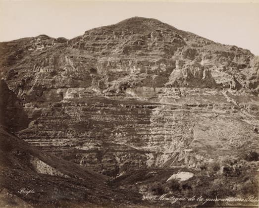 RECORD DATE NOT STATED View of the Mount of Temptation near Jericho, Montagne de la quarantaine (Palestine), photograph, Part of Travel Album with photos of Jerusalem and other biblical places., number: ... , negative number: 344 , photographer: Maison Bonfils, Jericho, c. 1867 - c. 1895, Palestine, West Bank, pasteboard, paper, albumen print, height 225 mm x width 281 mm, mountains, A wide view of a rocky hillside with layered strata and rough textured rock faces, showing horizontal ledges, small caves and recesses, and areas of lighter colored rock against darker surfaces, with a foreground of sloping grassy terrain and patches of exposed soil, and sparse low vegetation scattered across the lower slopes. Copyright Penta Springs LCK260531_03380