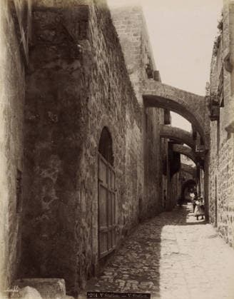 RECORD DATE NOT STATED Fifth Station of the Cross on the Via Dolorosa in Jerusalem, V Station. - V Station., photograph, Part of Travel Album with Photos of Jerusalem and Other Biblical Places., negative number: 264 , photographer: Maison Bonfils, Jerusalem, c. 1867 - c. 1895, pasteboard, paper, albumen print, height 282 mm x width 223 mm, street, stations of the cross, Via Dolorosa, A narrow cobblestone alley flanked by tall stone walls with multiple stone arches spanning overhead, wooden shutters and doors set into the walls, sunlight casting a bright strip along the center of the path while the sides remain in shadow, and several small figures farther down the alley including one adult and children wearing simple garments standing in the sunlit area. Copyright Penta Springs LCK260531_03383