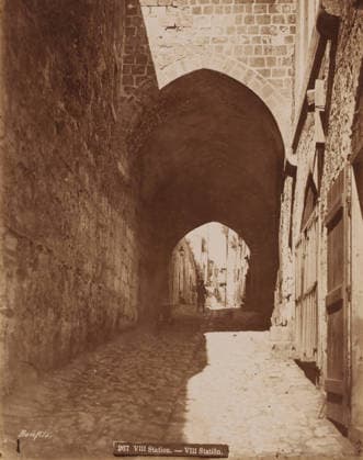 RECORD DATE NOT STATED VIII Station , eighth station in the Passion of Christ?, Jerusalem?, Palestine?, photograph, View of a gate, signature: Bonfils , inscription: 267 VIII Station. - VIII Station. , photographer: Maison Bonfils, c. 1880 - c. 1900, Maison Bonfils, photographer, pasteboard, paper, albumen print, height 277 mm, width 218 mm x width 344 mm, height 394 mm, A narrow cobblestone street runs beneath a large stone archway, with tall textured stone walls on either side; light falls on part of the right side of the street creating a bright patch on the ground while the left side remains in shadow, and a small silhouetted figure is visible in the distance under the arch with a few indistinct details of buildings and doorways beyond. Copyright Penta Springs LCK260531_17548