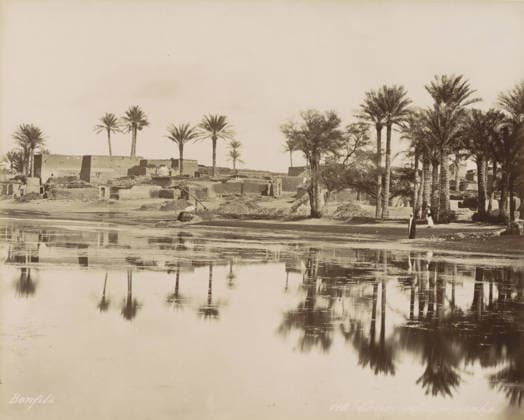 RECORD DATE NOT STATED Village with palm trees by a body of water near Cairo, Caire, village arabe, photograph, Part of Travel Album with 80 Photos of Egypt., negative number: 118 , photographer: Maison Bonfils, Cairo (city), c. 1870 - c. 1898, Ottoman Empire, North Africa, pasteboard, paper, albumen print, height 223 mm x width 280 mm, prospect of village, silhouette of village, trees: palm-tree, A wide sepia-toned riverside scene shows a shoreline with mud-brick buildings and low ruined structures partially obscured by clumps of vegetation. Tall palm trees rise along the bank, some clustered and some standing alone, their fronds splayed at the tops. A pair of figures in long robes stand near the trees by the water s edge. Reflections of the trees and shoreline Copyright Penta Springs LCK260531_18191