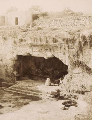 RECORD DATE NOT STATED Courtyard of the Tombs of the Kings in Jerusalem, Tombeau des Rois, cour intà rieure -Jà rusalem, photograph, Part of Travel Album with photos of Jerusalem and other biblical places., photographer: Maison Bonfils, Jerusalem, c. 1867 - c. 1895, pasteboard, paper, albumen print, height 223 mm x width 282 mm, grave, tomb, A weathered stone cliff face with a dark cave opening sits beneath a ledge carved with decorative rectangular and circular motifs, above which a rough terrace of grass and low vegetation gives way to a dry stone wall; to the left on the terrace a small square stone structure with an open doorway and nearby arched recesses is visible, and scattered stones and a short stepped platform lead down from the cave entrance to a flat, Copyright Penta Springs LCK260531_18257