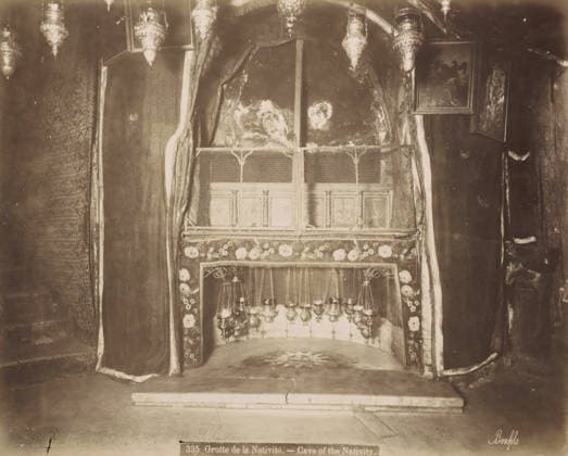 RECORD DATE NOT STATED Grotte de la Nativite.- Cave of the Nativity., Nativity Cave in the Church of the Nativity in Bethlehem, photograph, Part of Travel Album with Photos of Jerusalem and Other Biblical Places., negative number: 335 , photographer: Maison Bonfils, Bethlehem (Israel), c. 1867 - c. 1895, Palestine, pasteboard, paper, albumen print, height 225 mm x width 283 mm, interior of church, cave, grotto, A sepia-toned interior scene showing a small recessed alcove framed by heavy draped curtains with patterned borders and floral trim, a decorative metal grille across the upper front of the recess, hanging ornate lamps suspended inside the lower portion, a stone floor with a star-like design in front of the alcove, textured walls and framed pictures mounte Copyright Penta Springs LCK260531_18259