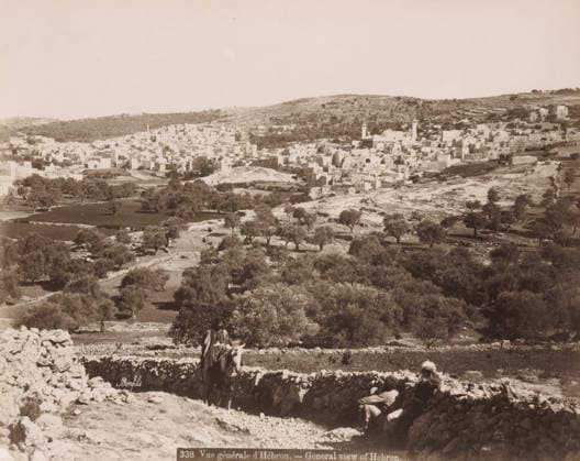 RECORD DATE NOT STATED General view of Hebron.- General view of Hebron., View of Hebron, photograph, Part of Travel Album with photos of Jerusalem and other biblical places., negative number: 338 , photographer: Maison Bonfils, Hebron, c. 1867 - c. 1895, Palestine, West Bank, pasteboard, paper, albumen print, height 222 mm x width 279 mm, prospect of city, town panorama, silhouette of city, A wide sepia-toned landscape showing a hillside town with densely packed light-colored stone buildings and a few taller tower-like structures set against rolling hills, with scattered trees and terraced fields in the foreground; a dry stone wall runs across the lower part of the scene, where two figures sit and one figure rides a donkey near the wall, and further down the slo Copyright Penta Springs LCK260531_18261